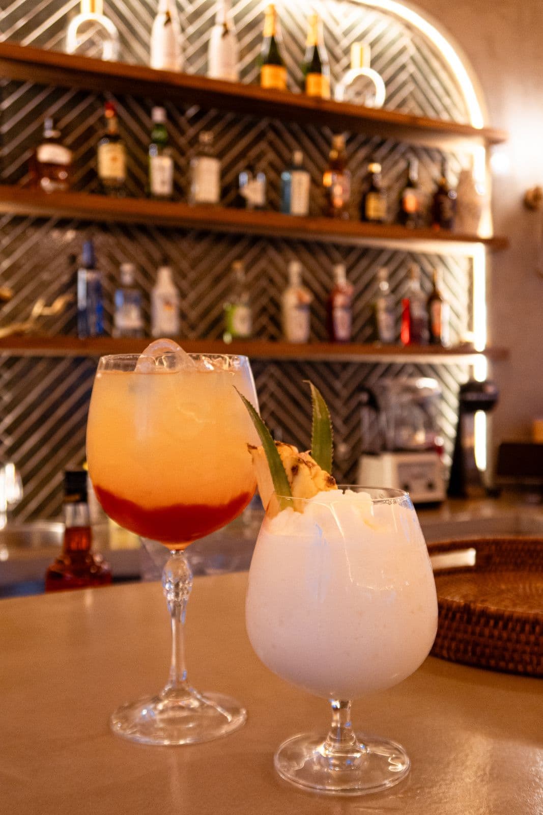 Zwei Cocktails auf dem Tresen, einer orangerot mit Eis, einer cremig weiß mit Ananas und Minze.