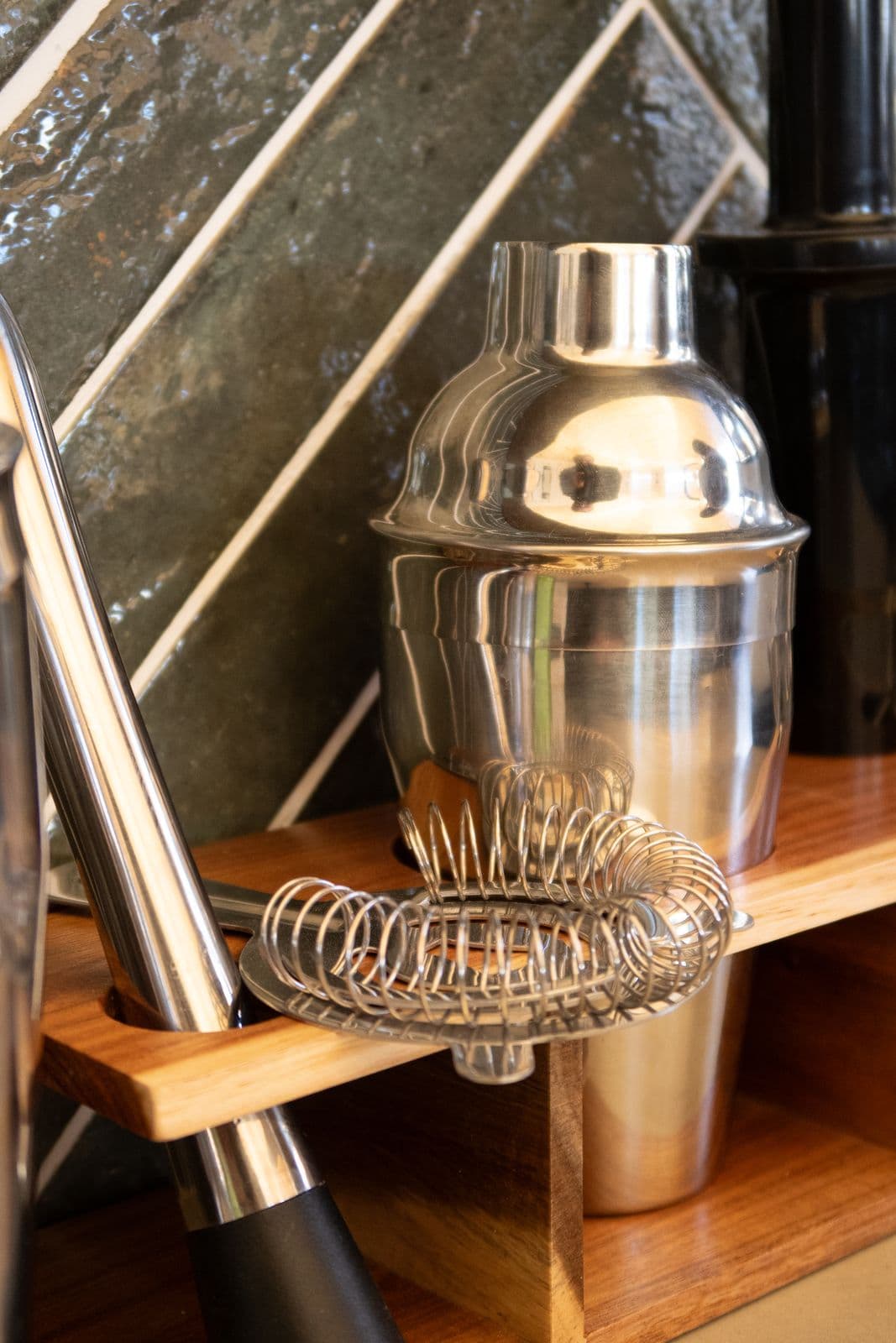 Cocktail-Shaker und Barsieb aus Edelstahl vor der grün gefliesten Wand.