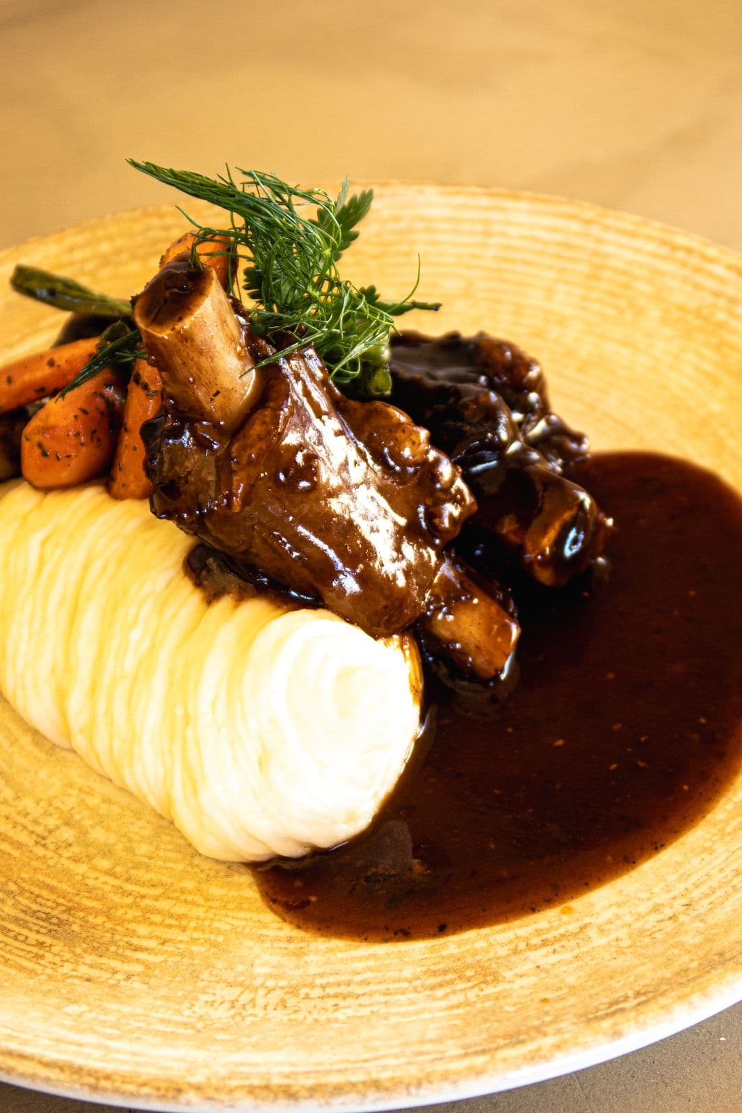Geschmortes Fleisch am Knochen mit Kartoffelpüree, dunkler Sauce, Karotten und frischen Kräutern.