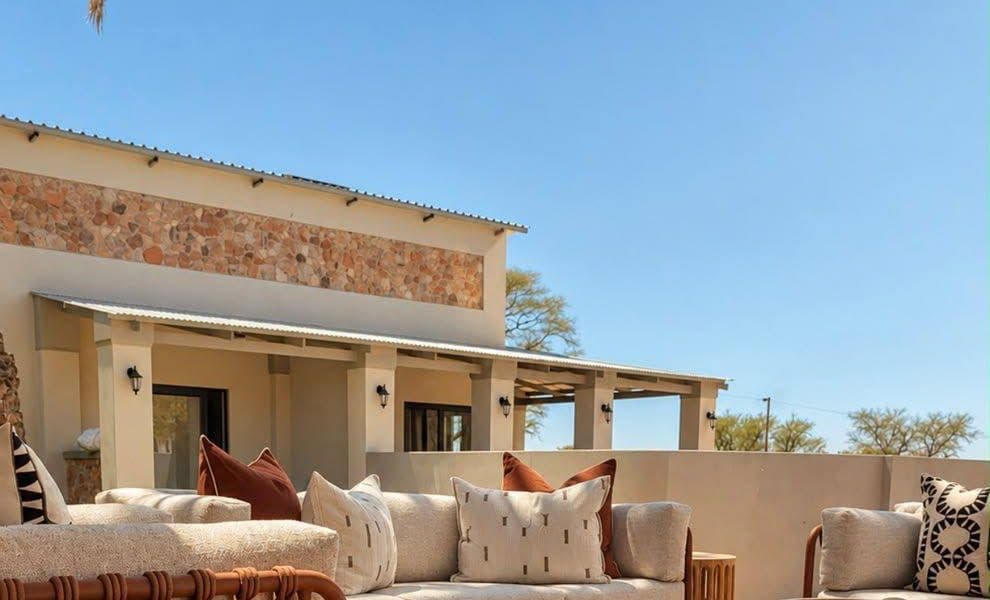 Lounge-Deck mit Sofas und Holztischen vor dem Farmhaus mit Natursteinband und Veranda, dahinter Akazien.
