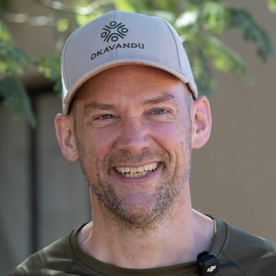 Torsten Herbert mit heller Okavandu-Kappe, lachend vor grünen Blättern.