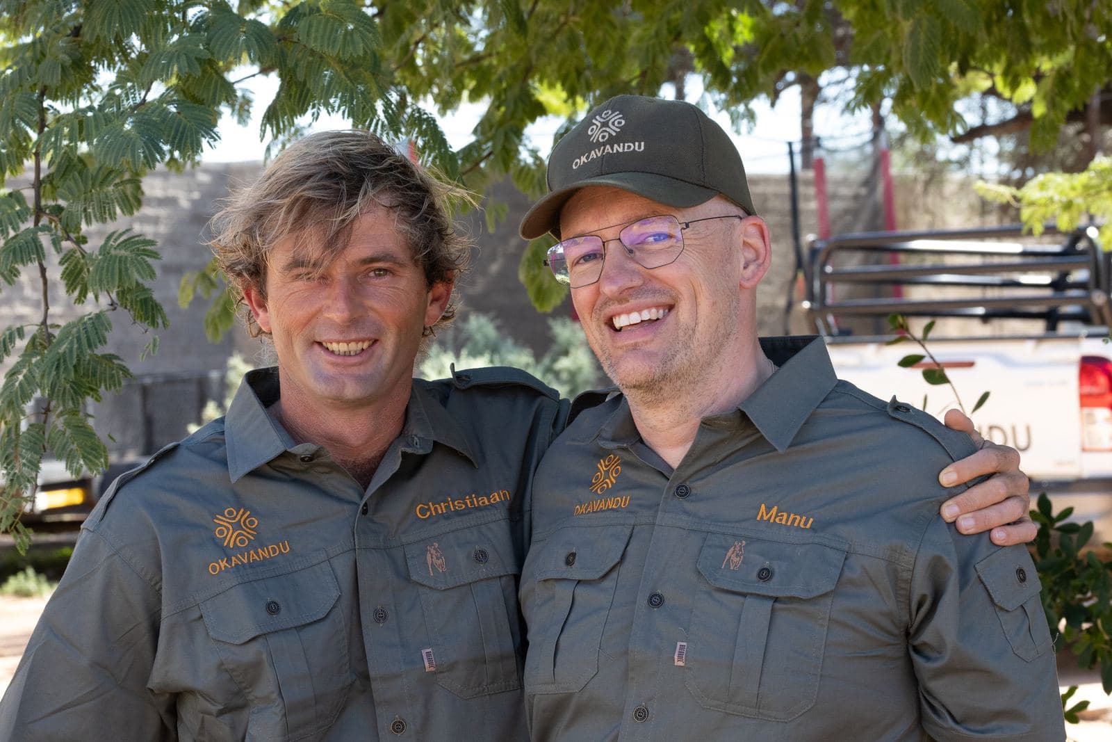 Chris und Manuel in Okavandu-Arbeitshemden, Arm in Arm und lachend.