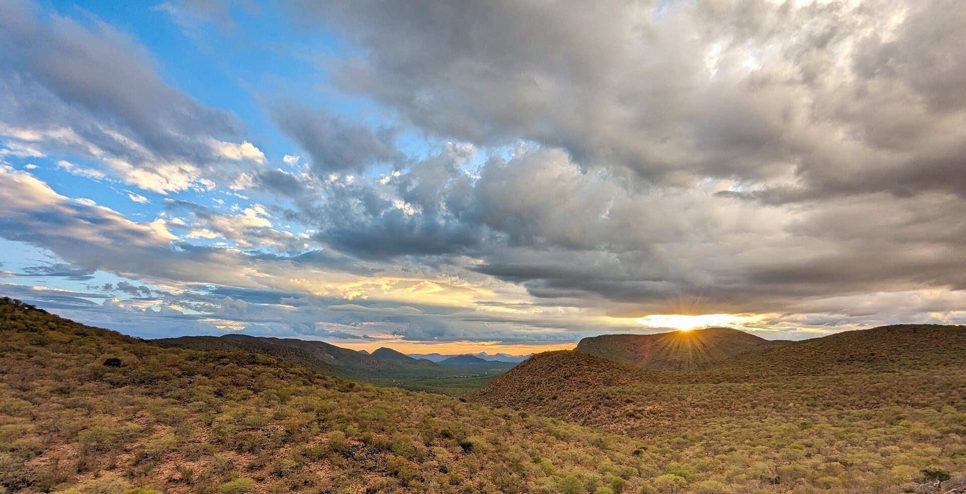 Die Abendsonne steht zwischen zwei Bergrücken des Erongo, darüber ziehen Wolken, im Vordergrund grasbewachsene Hänge.
