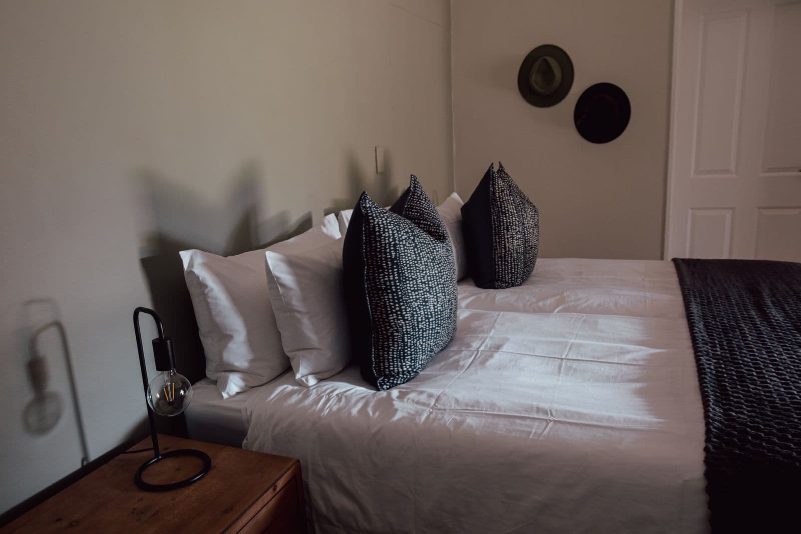 Schlafzimmer im Farmhaus mit Doppelbett, gemusterten Kissen und Nachttischlampe, an der Wand zwei Hüte.