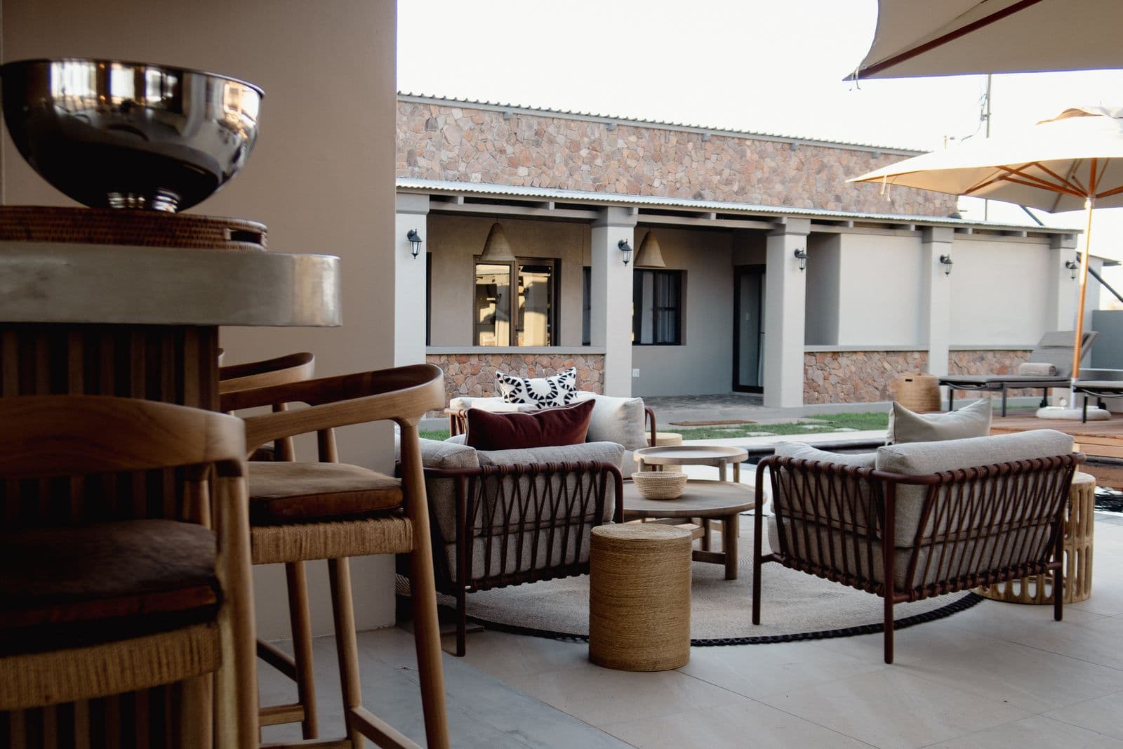 Lounge vor dem Zimmertrakt mit Rattansesseln und Rundteppich, vorn Barhocker.